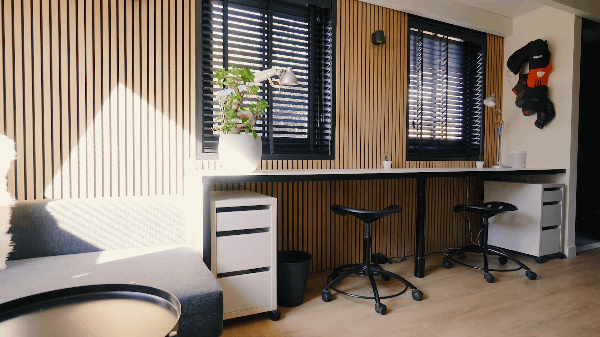 Modernes Büro mit nordischen Akustikholzpaneelen an den Wänden, ergonomischen Arbeitsplätzen, schwarzen Hockern, einer Topfpflanze und Sonnenlicht durch schwarze Jalousien.